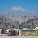 Pohled na dokončování jednotlivých vahadel. Panoráma mostu a jeřábů v zasněžené krajině doplňuje v pozadí výhled na obec Hrčava na české straně hranice.